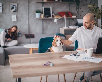 Ein Mann sitzt an einem Tisch und streichelt einen Beagle. Im Hintergrund sitzt eine Frau auf einem Sofa und schaut auf ihr Laptop. Der Raum ist modern und hell eingerichtet.